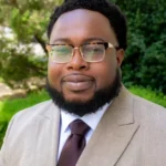 Portrait of a man wearing a beige herringbone suit, white shirt, brown tie, and glasses outdoors.