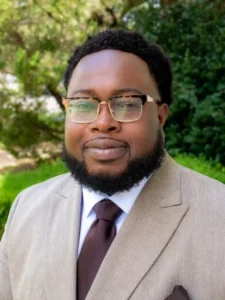 Portrait of a man wearing a beige herringbone suit, white shirt, brown tie, and glasses outdoors.